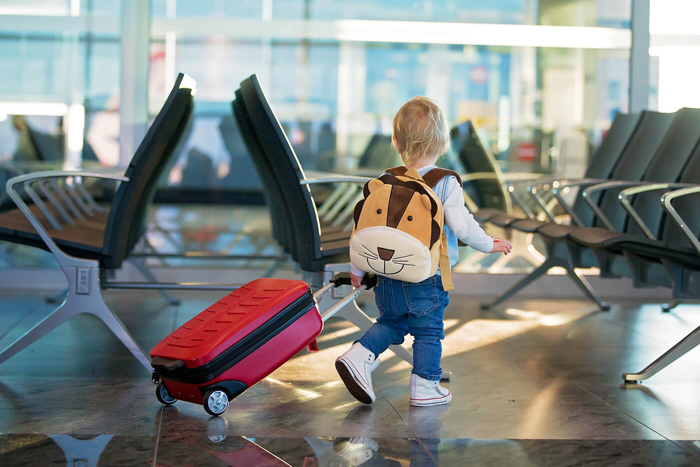 Toddler wearing lion backpack pulling red suitcase through airport terminal gate area ready for family trip