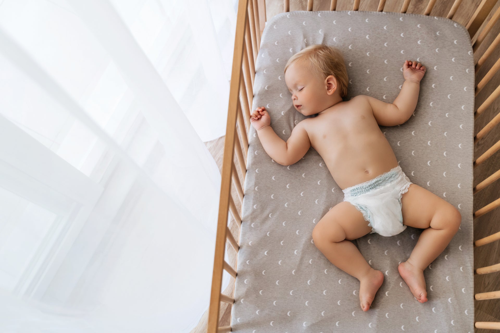 Peaceful baby sleeping in wooden travel crib with gray moon-patterned sheet during naptime at travel accommodation