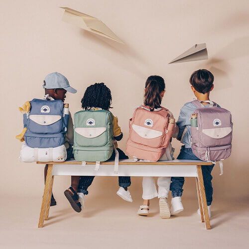 Back view of four children sitting on wooden bench wearing blue green pink and purple WeeWander backpacks.