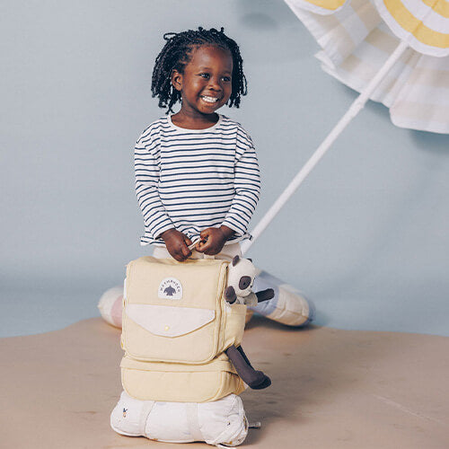 Smiling child wearing navy and white stripes perched on beige travel backpack with comfort toy demonstrating sturdy kid-friendly luggage.