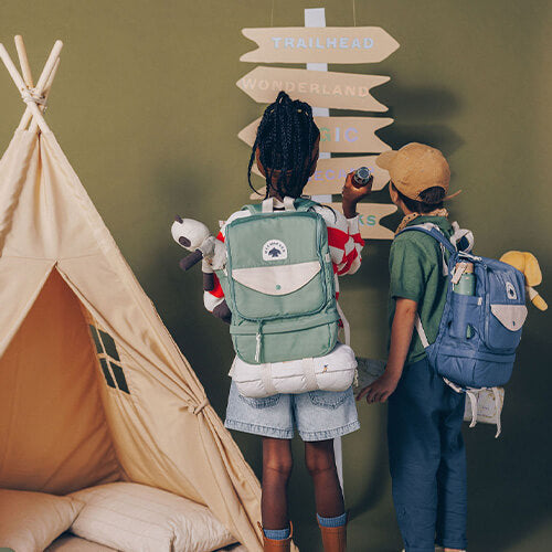 Back view of three children wearing green pink and blue backpacks looking up at wooden directional sign post deciding travel adventure path.