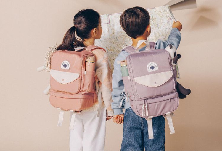 Back view of girl and boy wearing pink and purple WeeWander backpacks with water bottles studying large wall map for adventure planning.